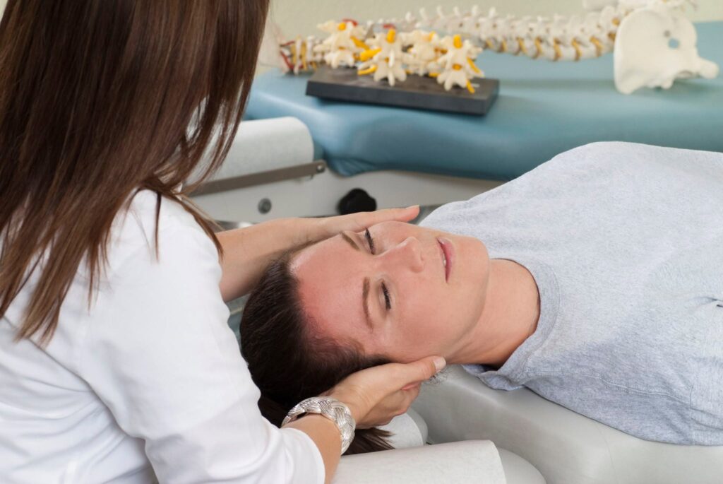 A chiropractor gently adjusting a patient's neck and head while the patient lies on a treatment table, with a spinal model in the background