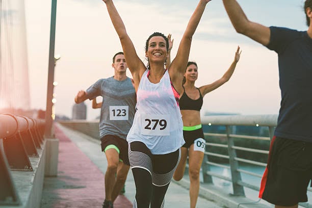 Woman wearing race bib 279 celebrates with arms raised while running a race on a bridge at sunset with other competitors