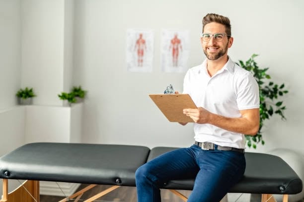 Smiling male physical therapist holding a clipboard, seated on a treatment table in a clinic with anatomy charts on the wall