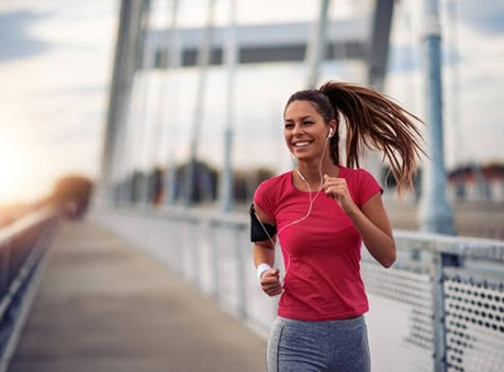 Woman jogging on a bridge, smiling, wearing earbuds and an armband phone holder