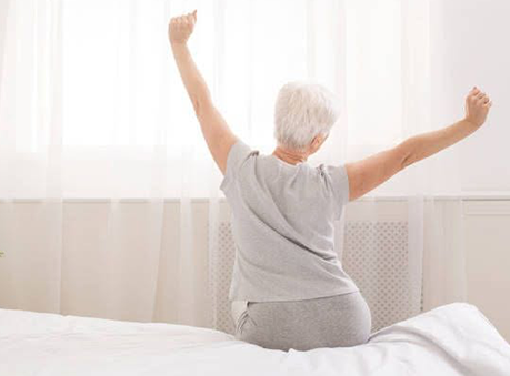 Older adult sitting on the edge of a bed, stretching arms overhead after waking up