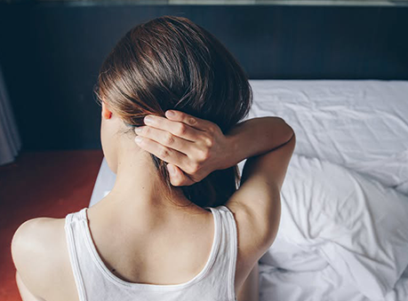 Woman sitting on a bed, seen from behind, holding the back of her neck in apparent pain or discomfort